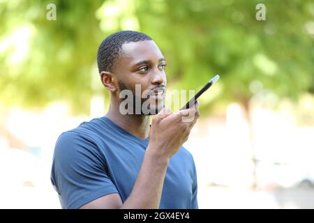 Homme noir dictant un message audio sur un smartphone qui marche dans un parc Banque D'Images