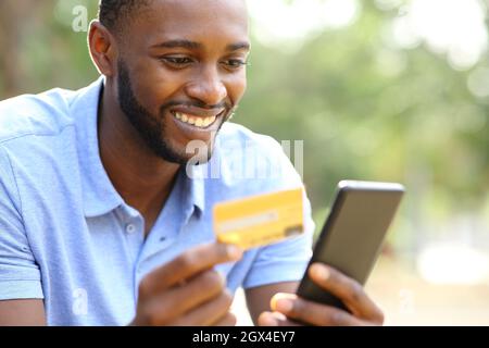 Joyeux homme noir achetant en ligne avec carte de crédit et smartphone dans un parc Banque D'Images