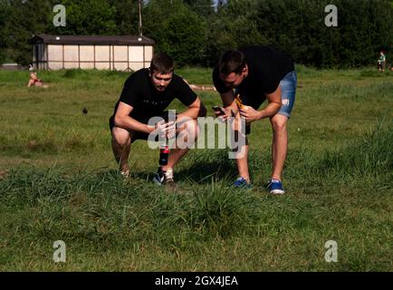 Deux jeunes hommes prennent des photos d'un gopher sauvage sur un téléphone cellulaire dans un pré avec des gens se reposant au milieu de l'herbe verte en été. Banque D'Images