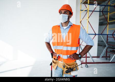 Portrait d'homme de course mixte constructeur en vêtements de travail et casque de sécurité portant un masque médical, gros plan Banque D'Images