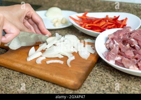 Gros plan des mains des femmes coupant des oignons blancs sur une planche à découper dans la cuisine, à côté des assiettes avec des morceaux de viande de porc, poivron rouge doux Banque D'Images