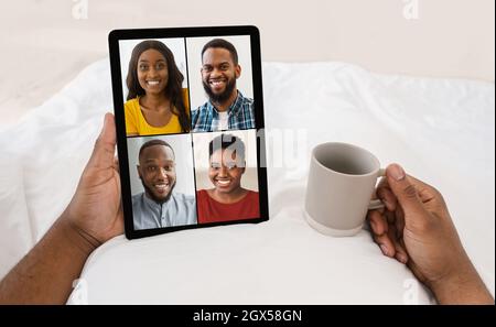 Un homme noir méconnaissable boit du café au lit, appelle de la famille ou des amis par vidéo, donne un coup de tête à différentes personnes Banque D'Images