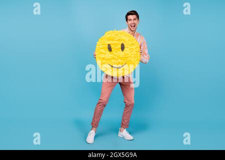 Photo pleine longueur de jeune gars heureux positif sourire tenir papier pinata émoticône isolé sur fond bleu de couleur Banque D'Images