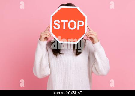 Portrait d'une femme inconnue avec des cheveux foncés tenant cachant son visage derrière le panneau d'arrêt rouge, interdiction, portant blanc pull de style décontracté. Studio d'intérieur isolé sur fond rose. Banque D'Images