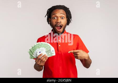 Homme étonné avec une bouche ouverte portant un T-shirt rouge de style décontracté, tenez beaucoup d'argent, pointant sur les billets en euros, choqué par la victoire soudaine. Prise de vue en studio isolée sur fond gris. Banque D'Images