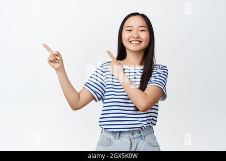 Bonne jolie fille asiatique montre la publicité, l'élève de l'adolescence pointant les doigts à gauche et sourire à la caméra, recommander le magasin, annoncer la vente ou la réduction Banque D'Images