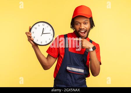 Livraison à temps. Homme de main optimiste dans les salopettes pointant à l'horloge, souriant à la caméra, professionnel et ponctuel service de réparation de maison, l'entretien. Studio d'intérieur isolé sur fond jaune Banque D'Images