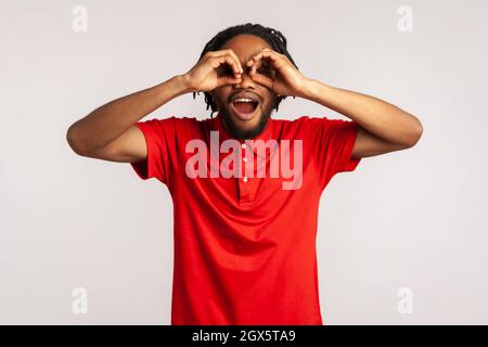 Émerveillé jeune homme adulte avec des dreadlocks portant un t-shirt rouge de style décontracté, regardant à travers les doigts en forme de jumelles et exprimant l'excitation. Prise de vue en studio isolée sur fond gris. Banque D'Images