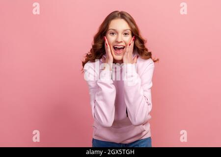 Portrait d'une adolescente heureuse surprise en sweat à capuche touchant le visage avec les mains et souriant sincèrement, humeur optimiste. Prise de vue en studio, isolée sur fond rose Banque D'Images