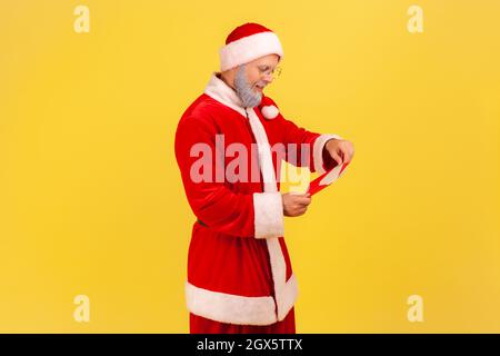 Vue latérale de l'homme âgé avec barbe grise en costume du père noël debout enveloppe rouge ouverte avec félicitations pour les vacances d'hiver, lire la lettre. Studio d'intérieur isolé sur fond jaune. Banque D'Images