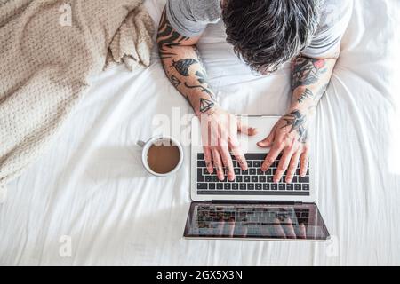 vue rognée de l'homme tatoué tapant sur un ordinateur portable dans le lit avec une tasse à café et des ordinateurs portables Banque D'Images