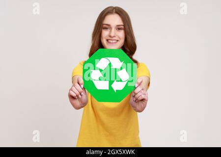 Jeune fille adolescente positive souriante avec cheveux bruns dans un style décontracté t-shirt jaune tenant le symbole vert de recyclage, triant les déchets, sauvant l'écologie. Prise de vue en studio isolée sur fond gris. Banque D'Images