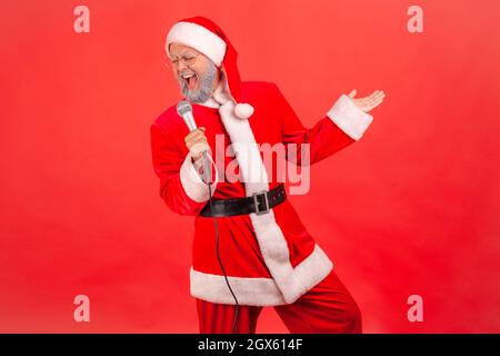 Joyeux homme âgé positif avec une barbe grise portant le costume du père noël debout avec un microphone dans les mains, chantant des chansons, célébrant Noël. Studio d'intérieur isolé sur fond rouge. Banque D'Images