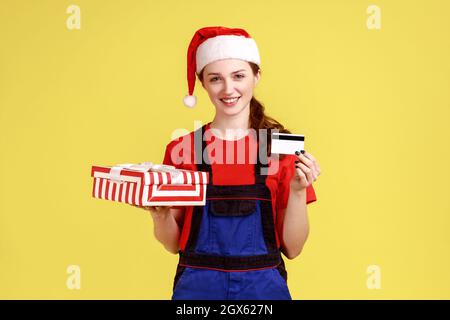 Bonne livraison femme tenant la boîte cadeau emballée et montrant la carte de crédit, bonus pour la livraison de la commande, portant la combinaison bleue et le chapeau du père noël. Studio d'intérieur isolé sur fond jaune. Banque D'Images