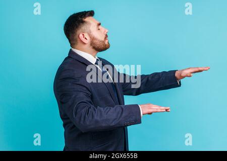 Vue latérale d'un homme aveugle malheureux portant un costume de style officiel debout avec les yeux fermés et l'air touchant pour trouver le chemin perdu, émulant la marche dans l'obscurité. Studio d'intérieur isolé sur fond bleu. Banque D'Images