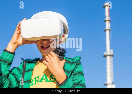 Enfant portant des lunettes de réalité virtuelle et tenant la caméra dans la main gauche, dans l'arrière-plan visible bleu ciel clair et relais de téléphonie mobile Banque D'Images