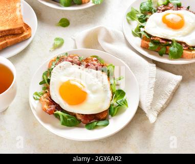 Des sandwichs ouverts avec des œufs frits, du bacon et des feuilles de légumes sur une assiette sur fond de pierre légère. Vue rapprochée Banque D'Images