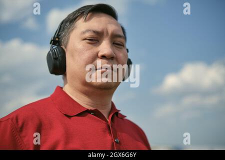 Homme asiatique d'âge moyen dans des écouteurs en plein air écoutant de la musique dans le ciel Banque D'Images