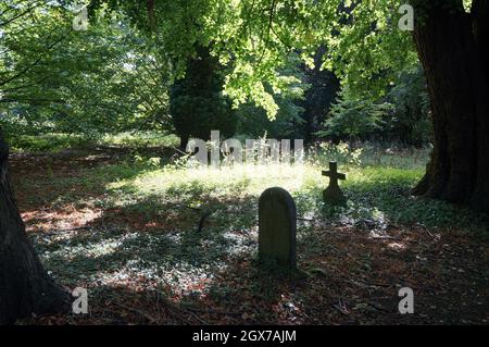 Tombe en pierre et une croix ou un crucifix dans un cimetière boisé avec la lumière du soleil filtrée sur l'herbe Banque D'Images