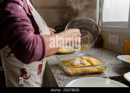 Vue latérale d'une femme qui verse la sauce Bechamel chaude sur du Cannelloni maison dans un plat à rôtir en verre Banque D'Images