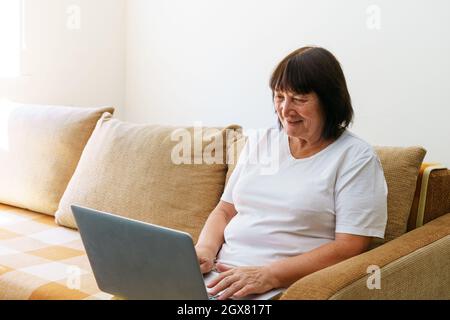 Une femme mûre surjoyeuse assise sur un canapé dans la salle de séjour, parlant sur un appel vidéo sur un ordinateur portable moderne, heureuse heureuse heureuse femme sénior saluant parler sur Internet à l'aide d'un ordinateur portable, concept de technologie Banque D'Images