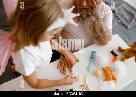 La fille peint les ongles de papa dans un vernis jaune vif à la table blanche dans la chambre Banque D'Images