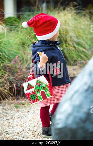 Petite fille habillée pour les promenades de Noël dans un parc avec sac de nouvel an.Chapeau rouge. Robe et collants, manteau bleu, sac en feutre en forme de maison d'hiver.Pas de face Banque D'Images