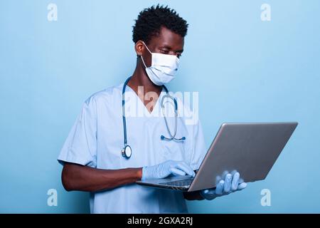 Homme infirmier avec uniforme tenant ordinateur portable sur un arrière-plan isolé dans le studio. Assistant médical avec masque facial et gants pour la protection du coronavirus tout en regardant l'appareil moderne en main Banque D'Images