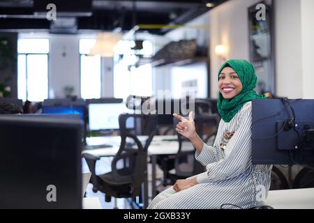 Portrait d'une femme africaine musulmane de développement de logiciels avec hijab vert assis sur le bureau de démarrage de plan ouvert moderne Banque D'Images