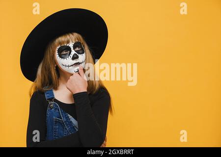 Petite fille réfléchie avec Halloween effrayant masque de maquillage réfléchit sur la décision, pensant à quelque chose, posant isolé sur fond jaune de couleur Banque D'Images
