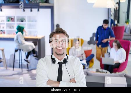 portrait d'un entrepreneur prospère avec des écouteurs autour de son cou et des collègues à l'arrière-plan au bureau de démarrage occupé Banque D'Images