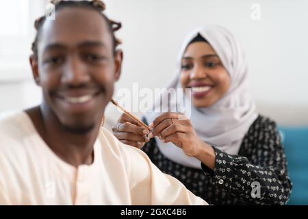 les jeunes couples musulmans afro ont un moment romantique à la maison tandis que la femme fait la coiffure à la femme de mari portant des vêtements traditionnels de hijab islamique soudanais Banque D'Images