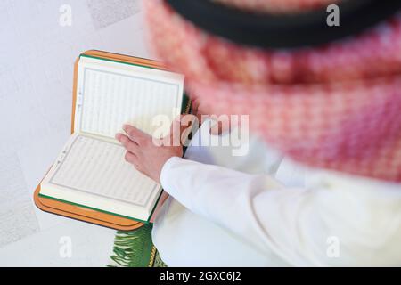 Jeune musulman arabe dans des vêtements traditionnels lisant le livre Saint Coran sur le tapis priant avant le dîner iftar pendant un festin de ramadan à la maison Banque D'Images