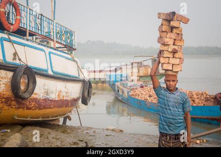 INDE, BANGLADESH - 8 DÉCEMBRE 2015 : un jeune homme ethnique vêt de vêtements sales qui marche avec des pierres en briques au-dessus de la tête près de la rivière et des bateaux qui regardent est venu Banque D'Images