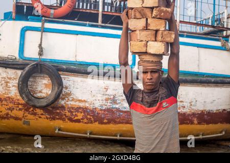 INDE, BANGLADESH - 8 DÉCEMBRE 2015 : un jeune homme ethnique vêt de vêtements sales qui marche avec des pierres en briques au-dessus de la tête près de la rivière et des bateaux qui regardent est venu Banque D'Images