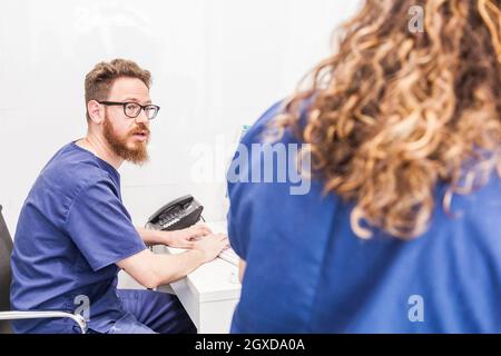 Homme barbu médic en uniforme et lunettes de vue dactylographiant sur le clavier tout en travaillant avec un médecin de culture à l'hôpital Banque D'Images