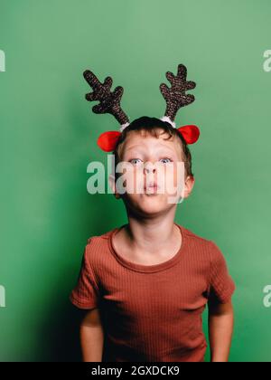 Mignon petit garçon dans les bois de cerf de Noël jante soufflant de l'air et sur fond vert et regardant l'appareil photo Banque D'Images