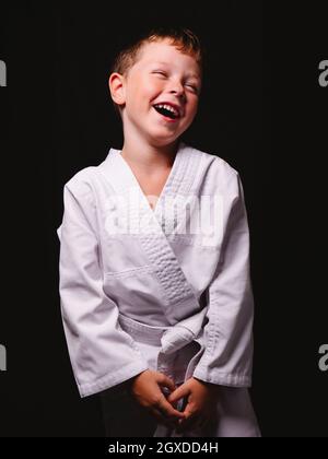 Mignon garçon dans un kimono karaté riant avec joie avec la bouche ouverte en studio sur fond noir Banque D'Images
