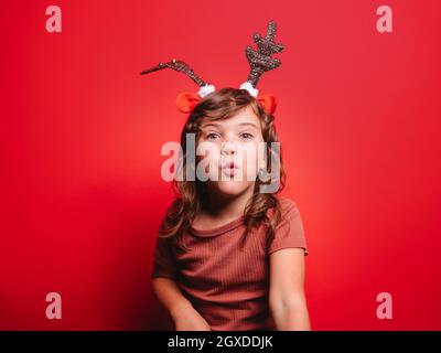 Joyeuse petite fille dans des vêtements décontractés et serre-tête cerf de fête soufflant baiser regardant l'appareil photo pendant la fête de Noël sur fond rouge Banque D'Images