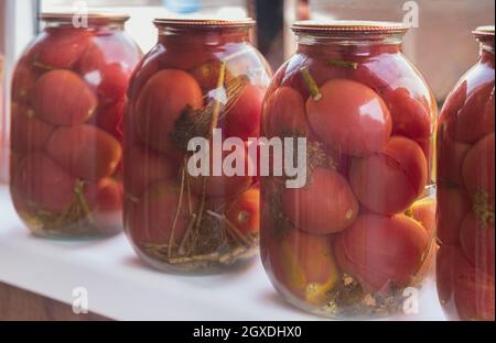 Accueil préservation: Grands pots en verre avec tomates rouges mûres marinées, fermés avec des couvercles en métal, se tiennent sur le rebord de la fenêtre. Vue avant, gros plan. Banque D'Images