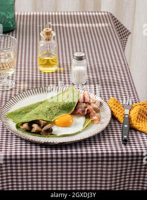Petit-déjeuner maison composé de crêpes aux épinards avec bacon, œufs et champignons servi sur une assiette blanche avec un saupoudreuse à l'huile et au sel sur une nappe à carreaux. Banque D'Images