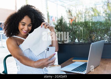 Une femme ethnique gaie avec une peau de vitiligo et un téléphone portable se promenant sur la promenade tout en regardant loin contre l'océan sous le ciel bleu Banque D'Images