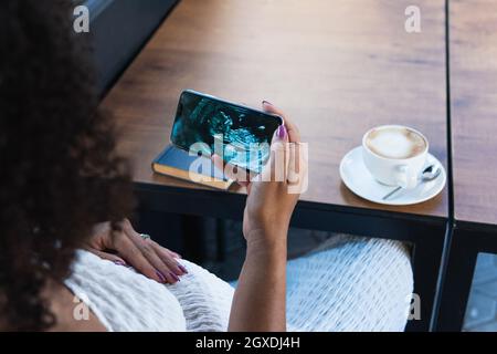 De dessus de la récolte femelle ethnique non reconnaissable assis à table avec une tasse de latte aromatique et livre et regardant bébé image d'ultrasons sur le téléphone cellulaire Banque D'Images