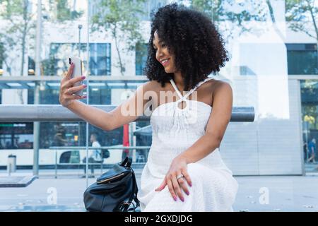 Jeune femme afro-américaine aux cheveux bouclés prenant le selfie avec un smartphone tout en étant assise sur le banc à l'arrêt des transports en commun Banque D'Images