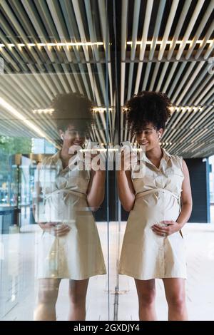 Agréable femme afro-américaine enceinte dans des écouteurs sans fil avec des messages texte sur les cheveux bouclés sur le téléphone mobile près du bâtiment en verre Banque D'Images