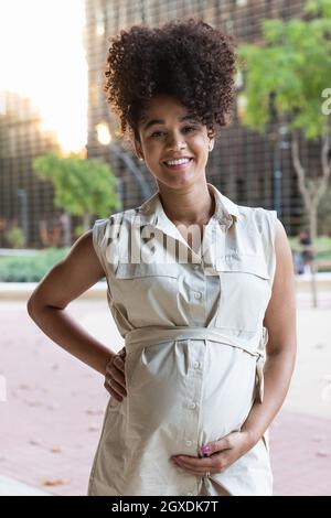 Femme afro-américaine enceinte positive en robe touchant le ventre et regardant la caméra Banque D'Images