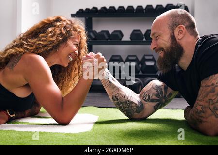 Vue latérale de la Sportswoman hispanique souriante avec les cheveux bouclés main de bodybuilder gai pendant le défi de lutte de bras contre les haltères dans la salle de gym Banque D'Images