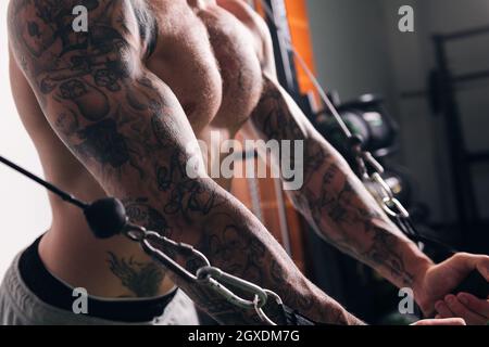 Vue latérale de la récolte sportif musclé concentré avec tatouages faire des exercices sur la machine de câble croisé dans la salle de gym avec des murs légers Banque D'Images