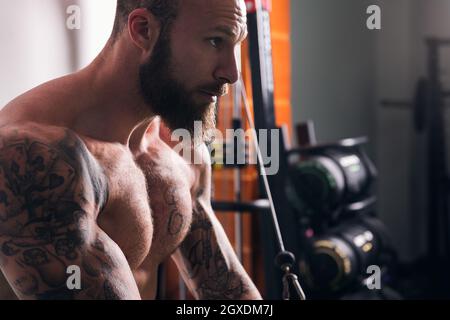 Vue latérale de la récolte sportif musclé concentré avec tatouages faire des exercices sur la machine de câble croisé dans la salle de gym avec des murs légers Banque D'Images