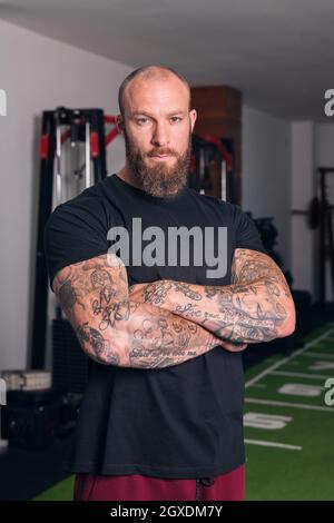 Sportif adulte fort avec barbe et tatouages sur les bras pliés regardant la caméra dans le gymnase Banque D'Images